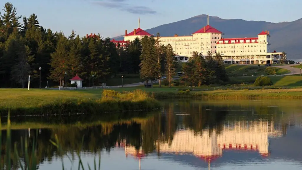 A view of the white and red Omni Mount Washington with a lake in front and mountains behind