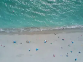 Aerial view of beach with clear turquoise water and white sand dotted with beach umbrellas, people, and chairs