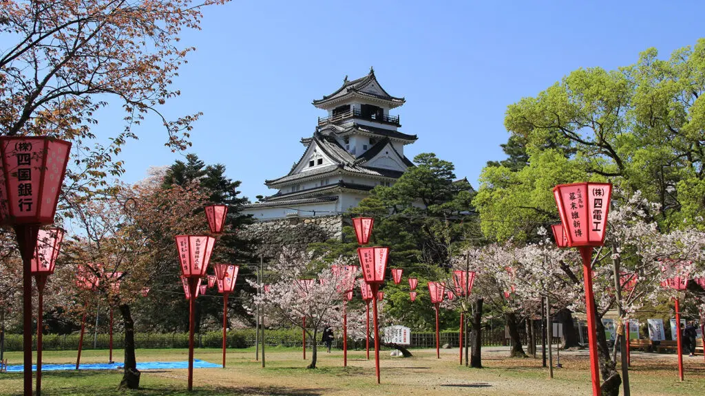 Ancient temple set behind green trees and blooming cherry blossoms, with Japanese lanterns in the forefront