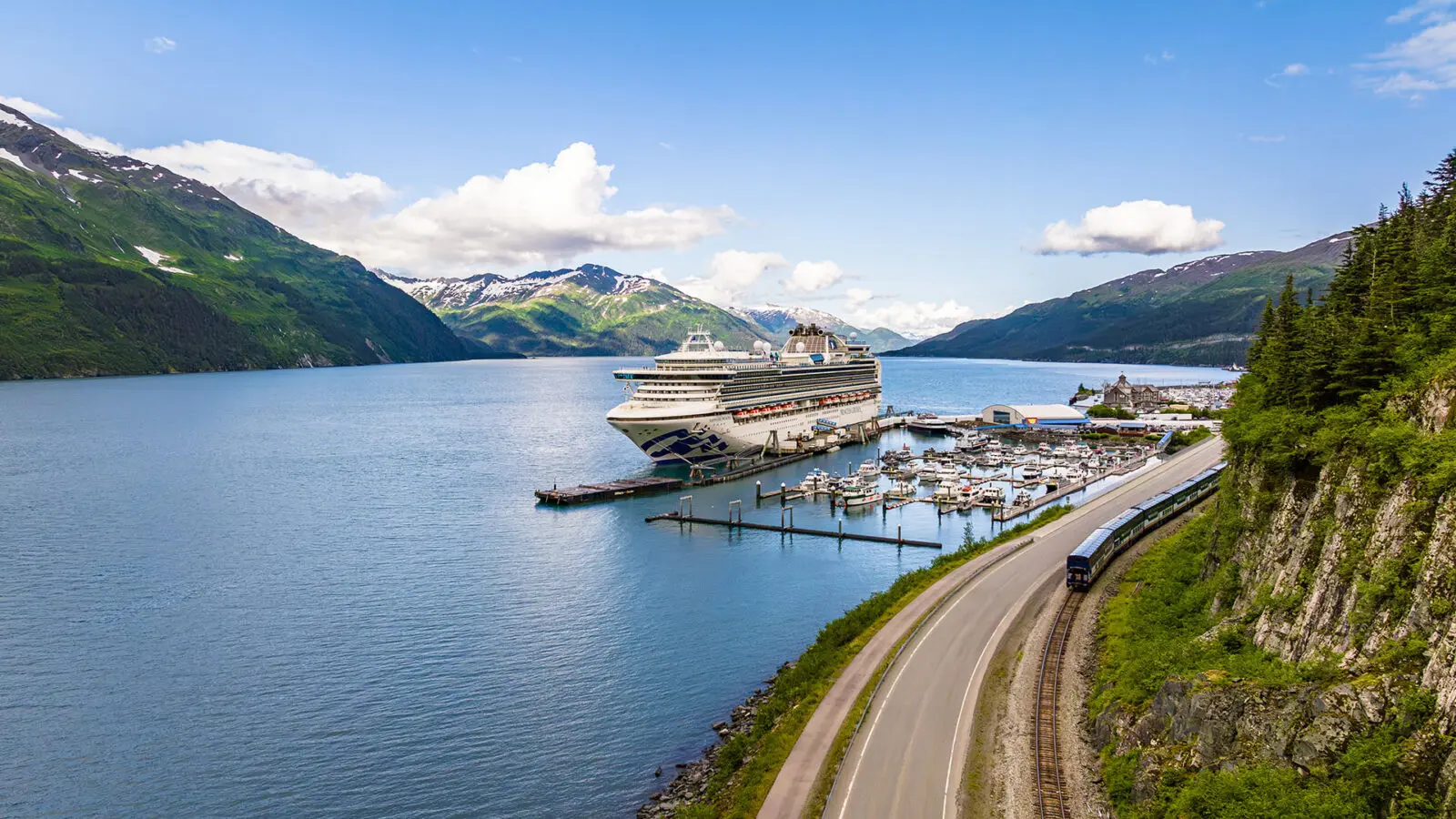 Cruise ship docked in bay alongside a bluff with a train going by and mountains in the background