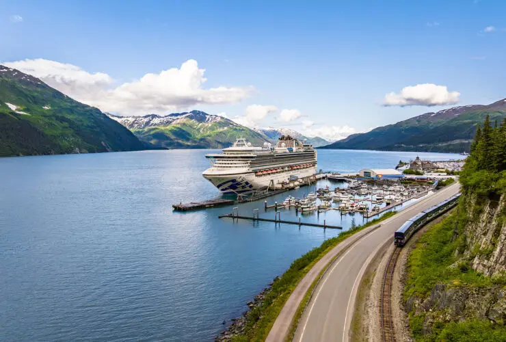 Cruise ship docked in bay alongside a bluff with a train going by and mountains in the background