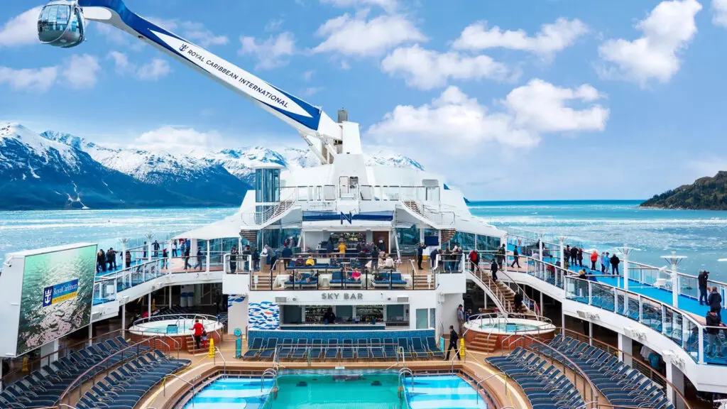 Cruise ship top deck with pool and large metal arm cantilevered over the edge of the ship, with snow-capped mountains in the background