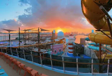 Cruise ship deck with water slides and a ropes course at sunset, with city skyline in background