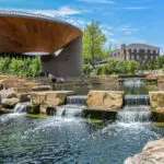 People hanging out at pond with tiered waterfall feature in park with large pavilion and buildings in background