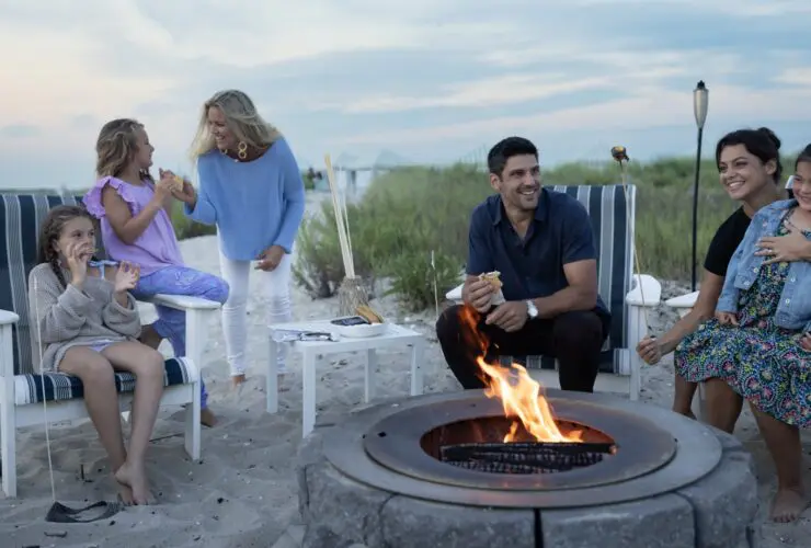 Adults and kids sitting around a firepit on the beach eating s'mores