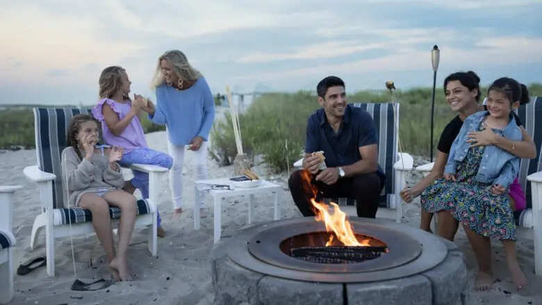 Adults and kids sitting around a firepit on the beach eating s'mores