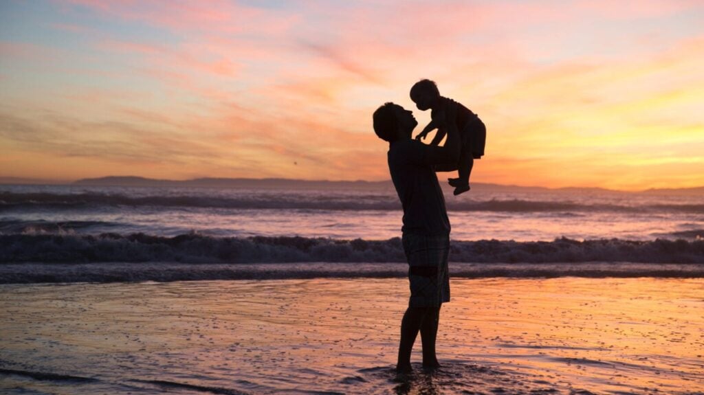 Adult lifting baby on beach at sunset