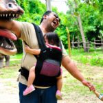 parent with young child and baby joking with dinosaur statue