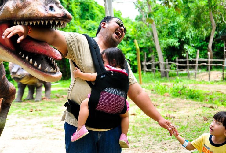 parent with young child and baby joking with dinosaur statue