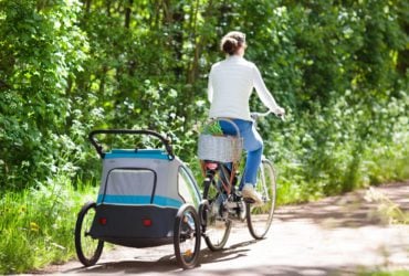 Young mother riding bicycle with baby bike trailer in sunny summer park. (Photo: Shutterstock)