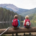 kids wearing kids backpacks sitting on a log overlooking a lake and forest