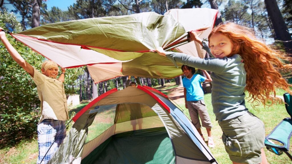 three kids working together to throw a rain tarp over the top of a tent while on a family camping trip