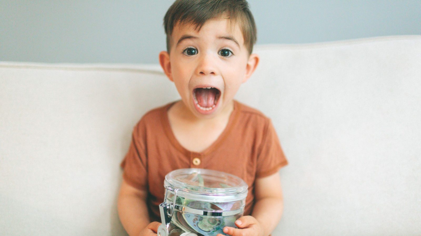 child sitting on couch making a surprised face and holding a jar of money; teaching kids about money