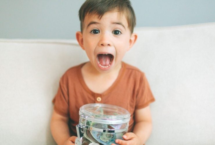 child sitting on couch making a surprised face and holding a jar of money; teaching kids about money