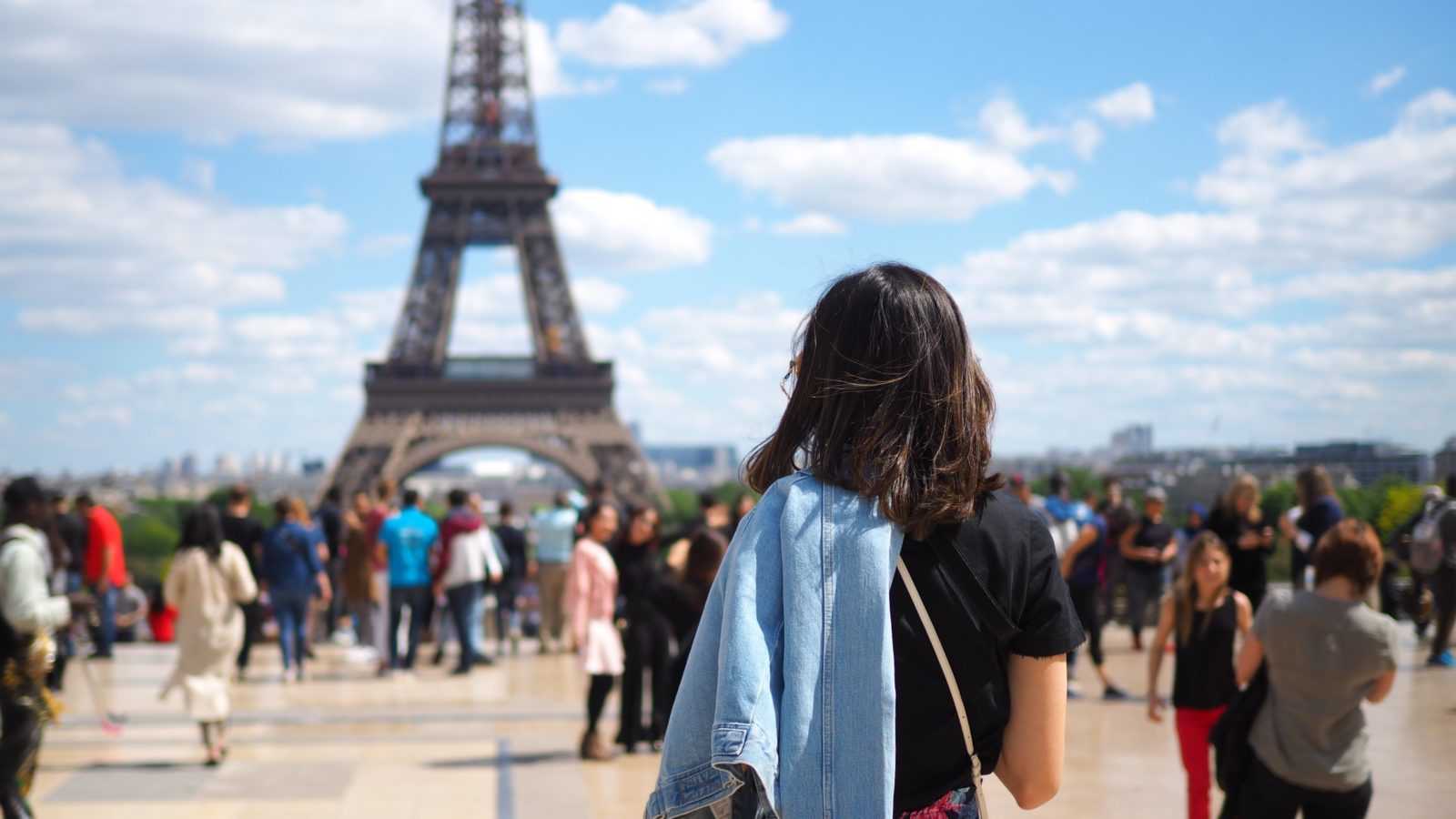 Woman in foreground looking out at a crowd of people walking and standing wearing a wide variety of outfits and shoes with the Eiffel Tower