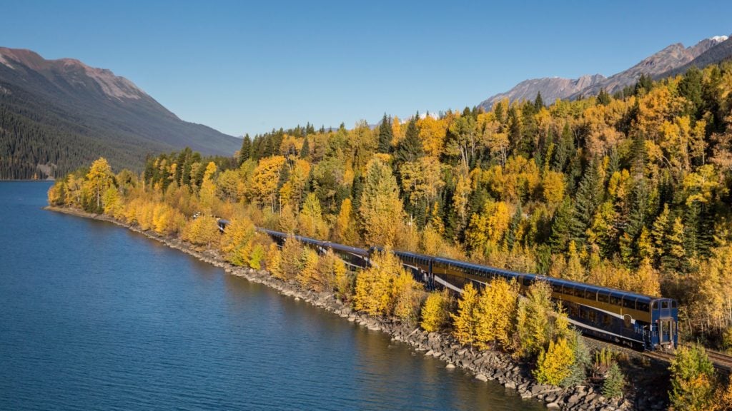 Kereta Rocky Mountaineer di sebelah air dan hutan musim gugur di jalur Journey through the Clouds