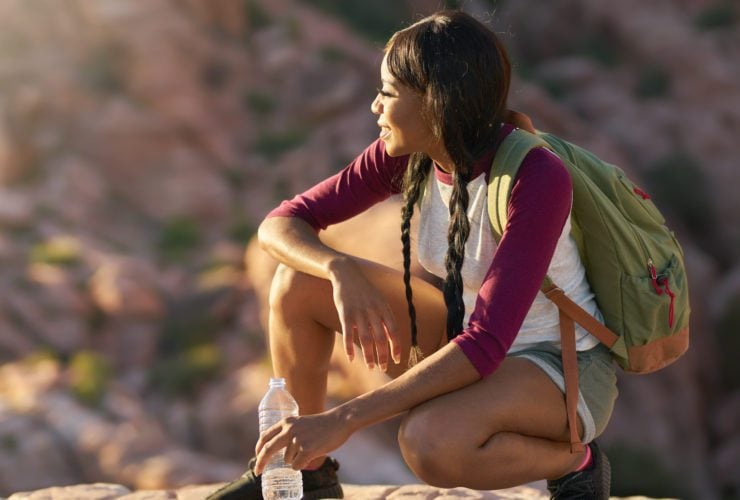 woman wearing travel backpack relaxing on a rock with a waterbottle at sunset