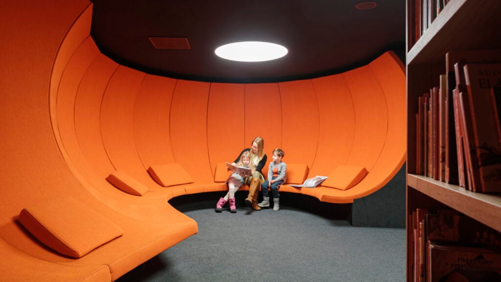 Orang dewasa dan anak-anak duduk di kursi tinggi oranye terang di Oodi di Helsinki