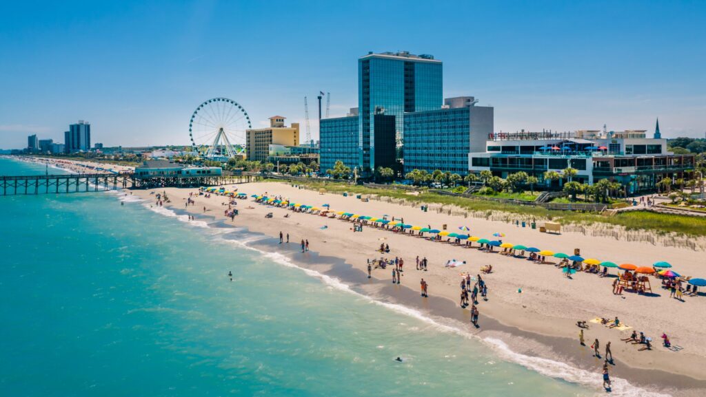 An aerial view of Myrtle Beach wiht lots of beach goers and umbrellas with hotels and the Ferris wheel in the background