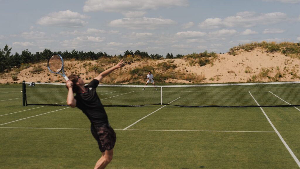 Two people playing tennis on a grass court with sand dunes behind them at Sand Valley resort