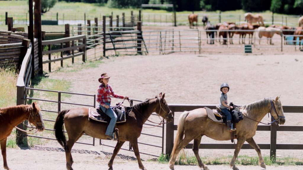 A child and adult rides horses by a corral