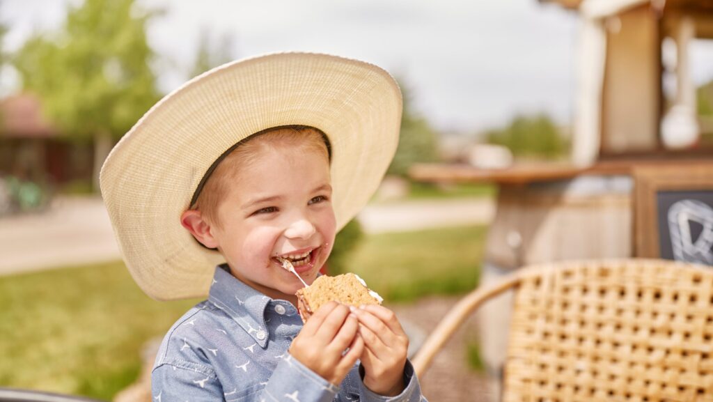 A little boy in a cowboy hat eating s'mores outside