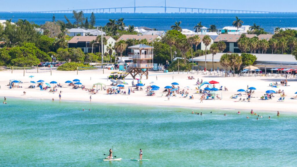 Umbrellas and chairs on the sand on Manatee Beach, with paddleboarders in the water and a lifeguard stand and the Sunshine Skyway behind