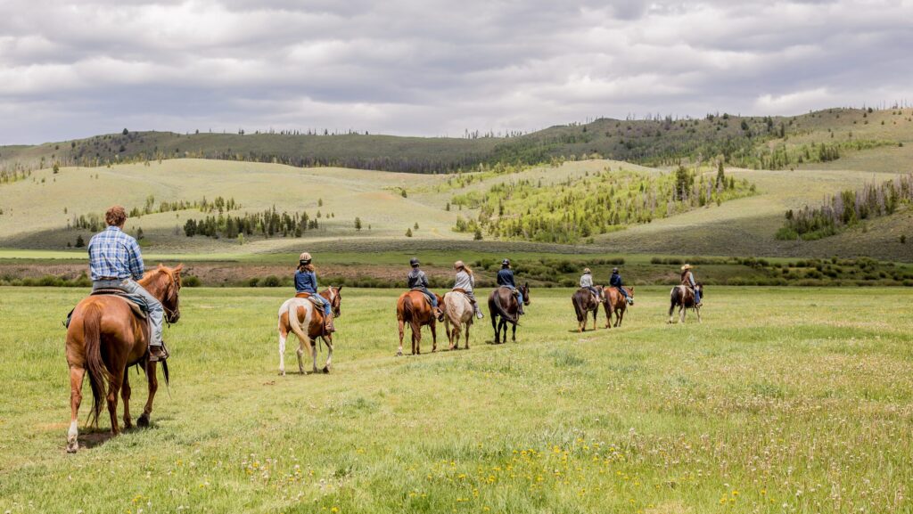 A group of people riding horses across a meadow with mountains in the distance at C Lazy U Ranch