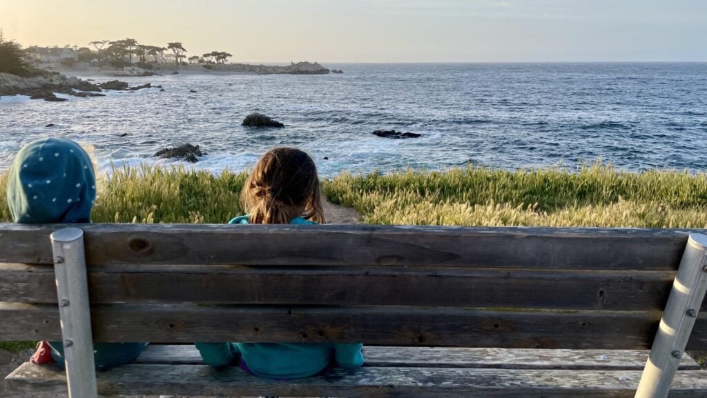 View of kids sitting on a bench along the walking path looking out at Lovers Point Beach in Monterey/Pacific Grove