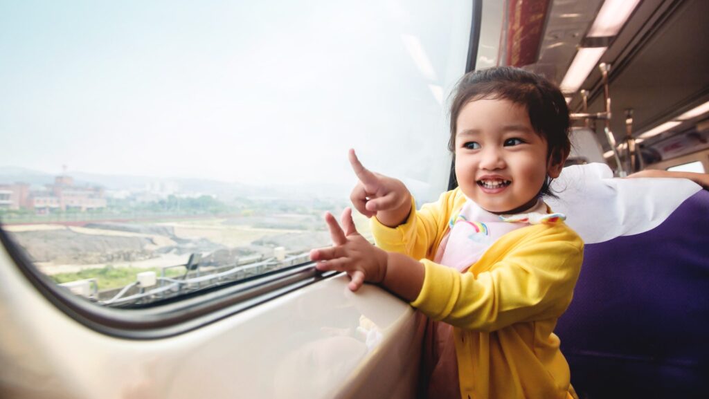 adorable child in a yellow jacket on a train looking out window while smiling and pointing