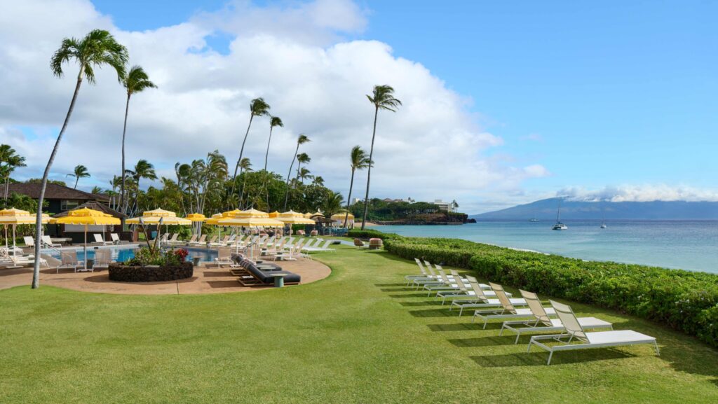 Lounge chairs lined up on the grass by a pool with yellow and white striped umbrellas overlooking the beach at Royal Lahaina Resort & Bungalows on Maui