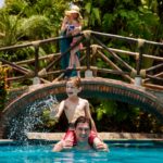 A mom and daughter squirting a squirt gun from a bridge at a dad and son in the pool
