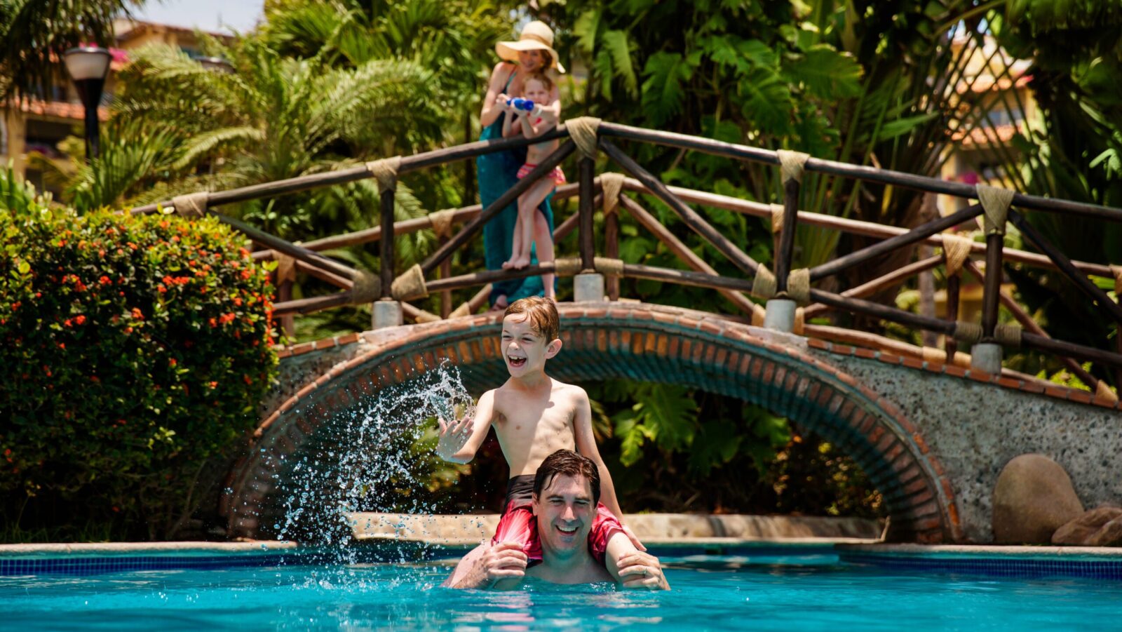 A mom and daughter squirting a squirt gun from a bridge at a dad and son in the pool