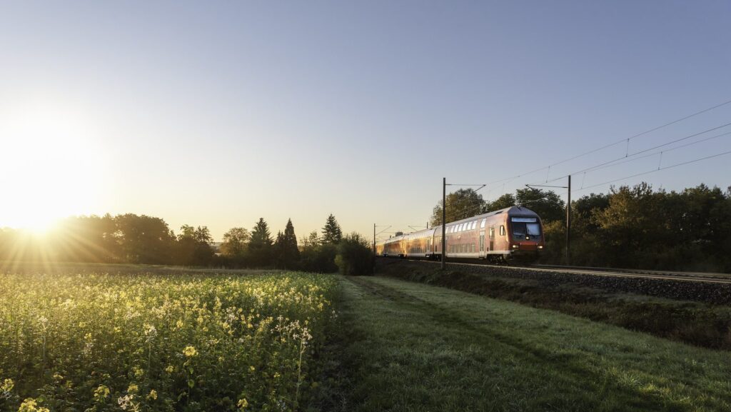 German Regional Train And Rapeseed Field At Sunrise. Passenger Train Traveling In Spring Scenery