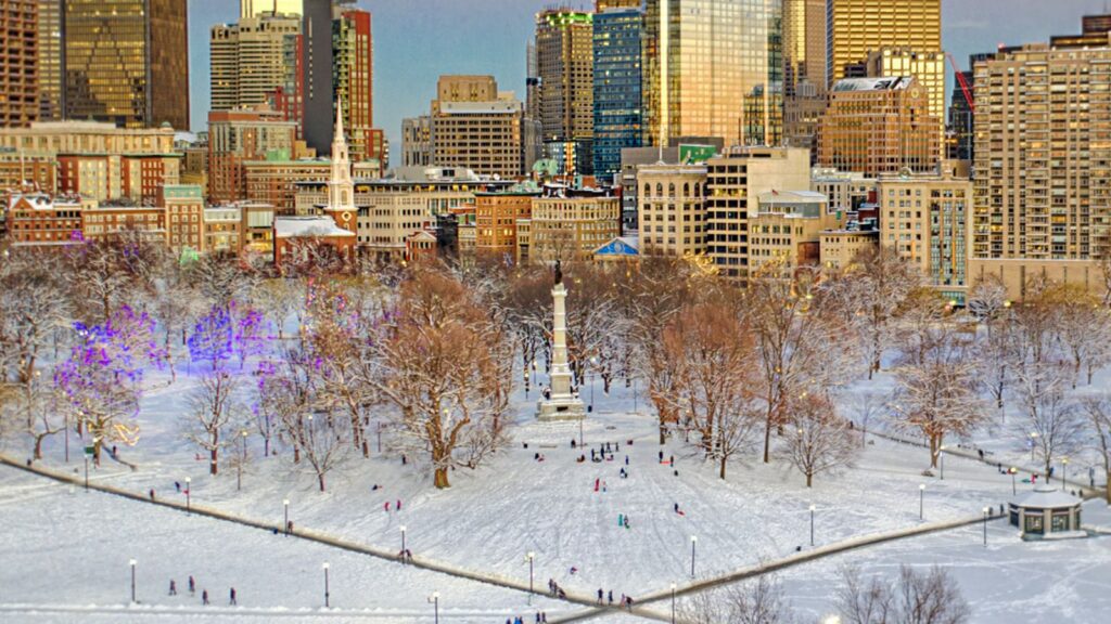 aerial view of Boston common covered in snow with people walking along the paths and the Boston skyline in the distance