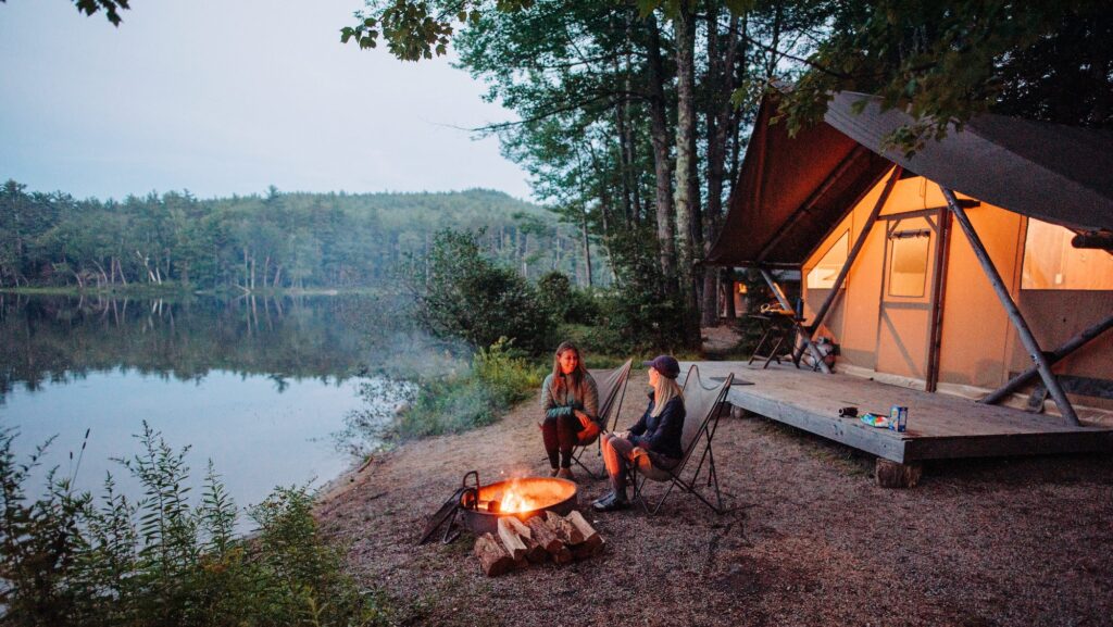 two people sitting by a campfire next to a glamping tent on a lake at a Huttopia resort in the U.S.