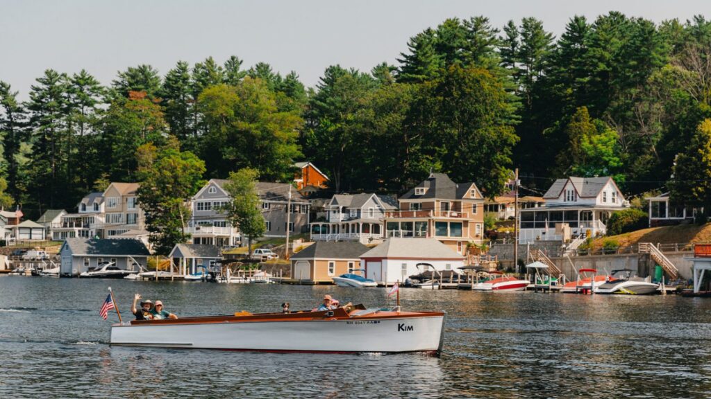 A wooden boat driving past houses lining Lake Winnipesaukee
