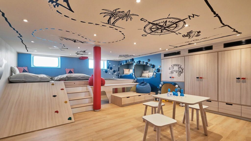 Nautical-themed children's club with compass and map art on ceiling, table with games, and ramp with bench and mini rock wall