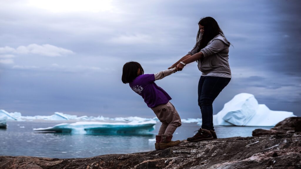 Woman holding young girl's hand and swaying her on rocky terrain with icebergs in the background