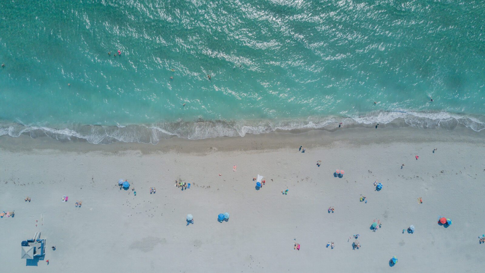 Aerial view of beach with clear turquoise water and white sand dotted with beach umbrellas, people, and chairs