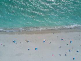 Aerial view of beach with clear turquoise water and white sand dotted with beach umbrellas, people, and chairs