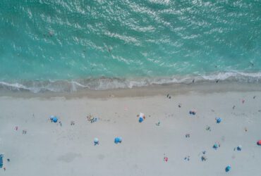 Aerial view of beach with clear turquoise water and white sand dotted with beach umbrellas, people, and chairs