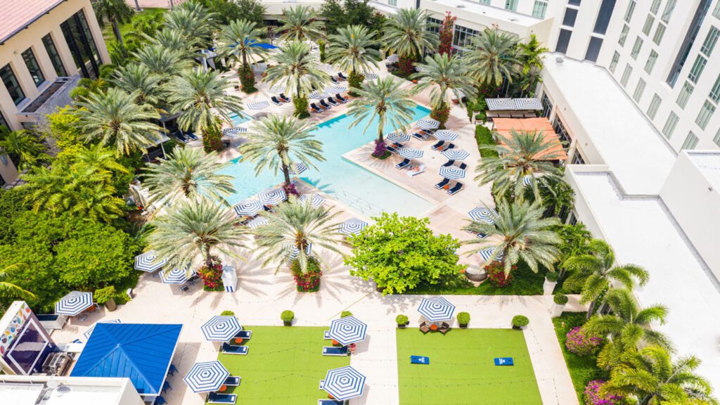 Aerial view of swimming pool and deck dotted with palm trees and umbrellas adjacent to a green lawn space