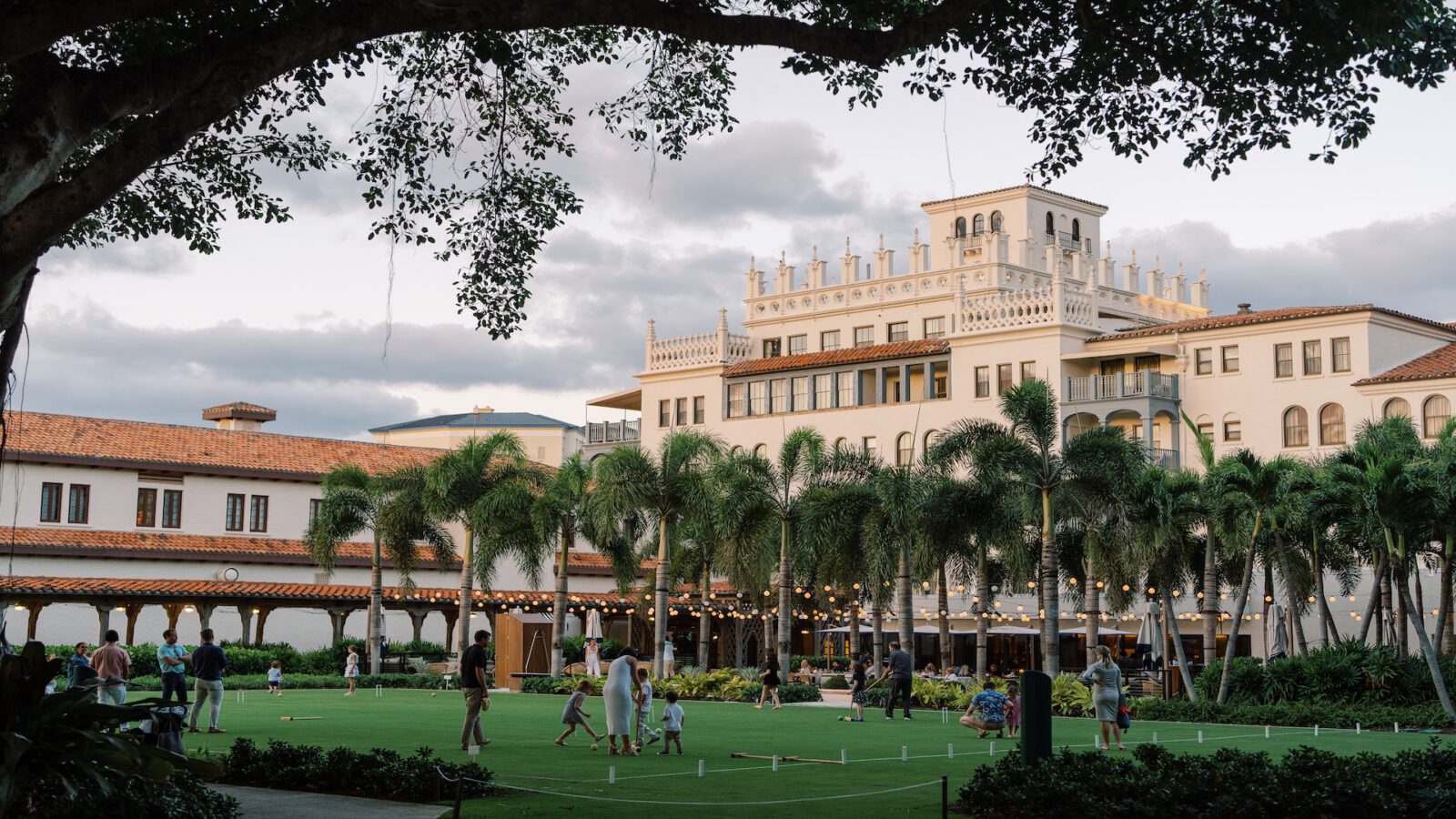 Palatial-style resort building at sunset with a large green lawn space in the forefront adorned by string lights and with families playing games