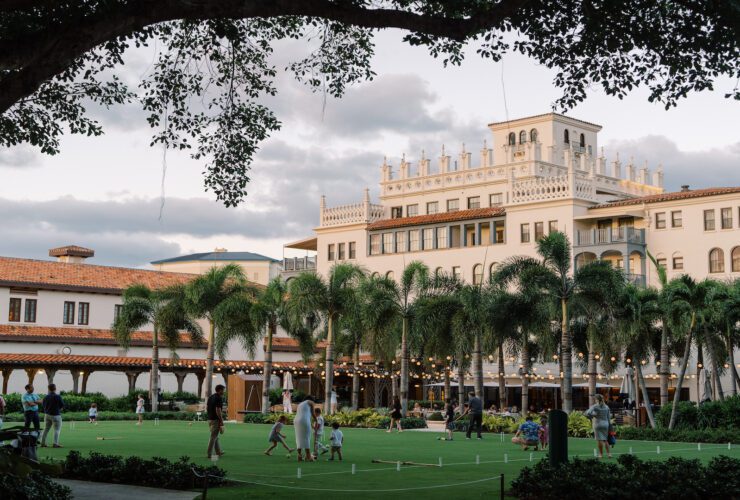 Palatial-style resort building at sunset with a large green lawn space in the forefront adorned by string lights and with families playing games