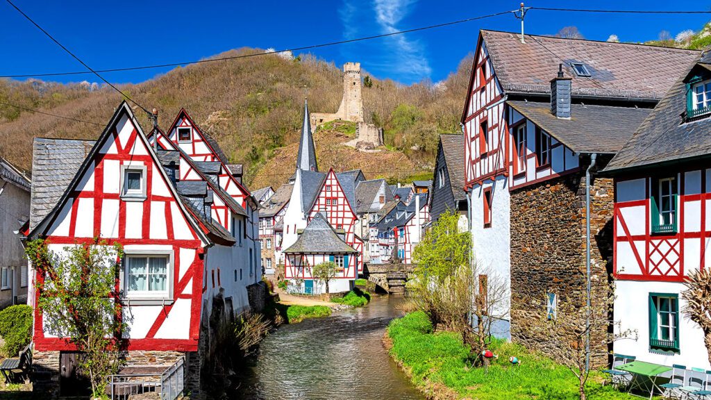 White half-timbered houses with red trim nestled in a small village along a stream, with a castle on a hill in the background