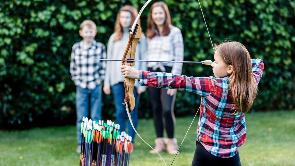 Girl preparing to shoot a bow and arrow with three other kids watching her in the background