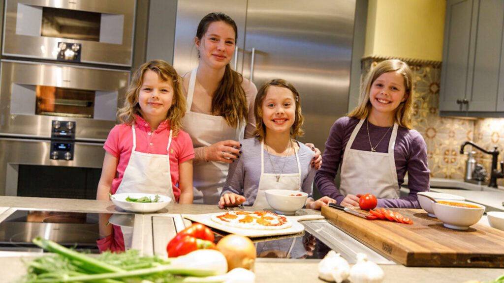 Three girls learning how to cook in a training kitchen, standing next to their instructor, with ingredients on the table