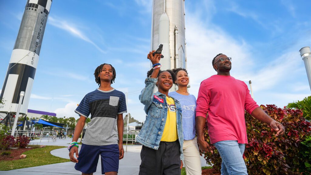 Two parents and their daughter and son walk outside complex with rocket structures in the background