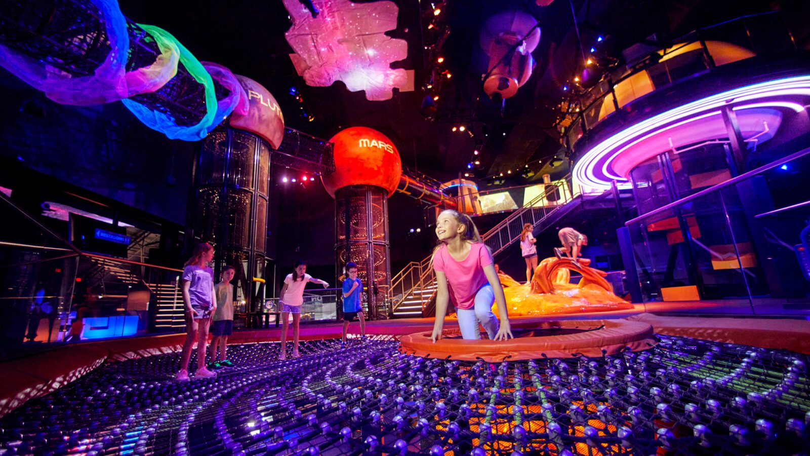 Kids play in dark, colorful space with planet structures hanging from the ceiling